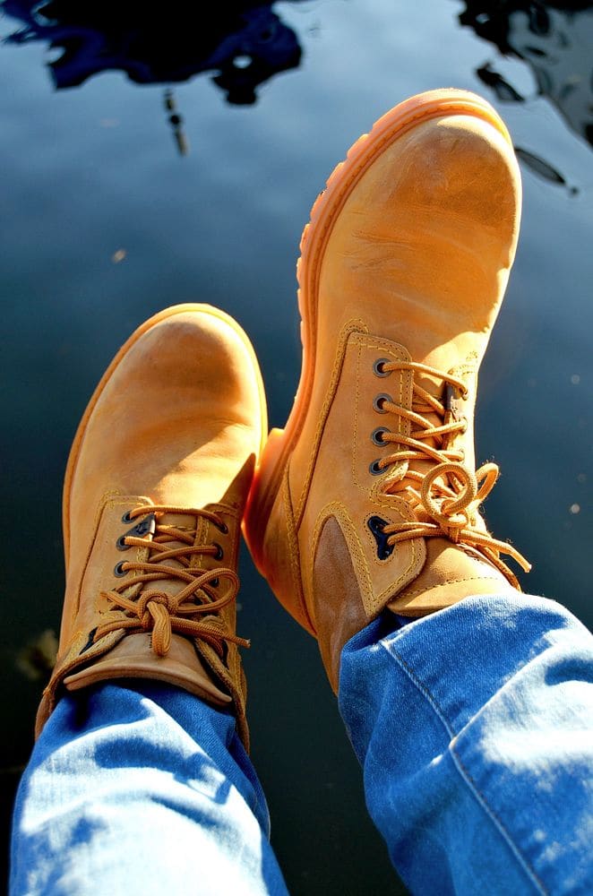 A pair of wheat-colored leather boots with long laces, crossed at the ankle and raised against a dark blue background, cuffed jeans above.