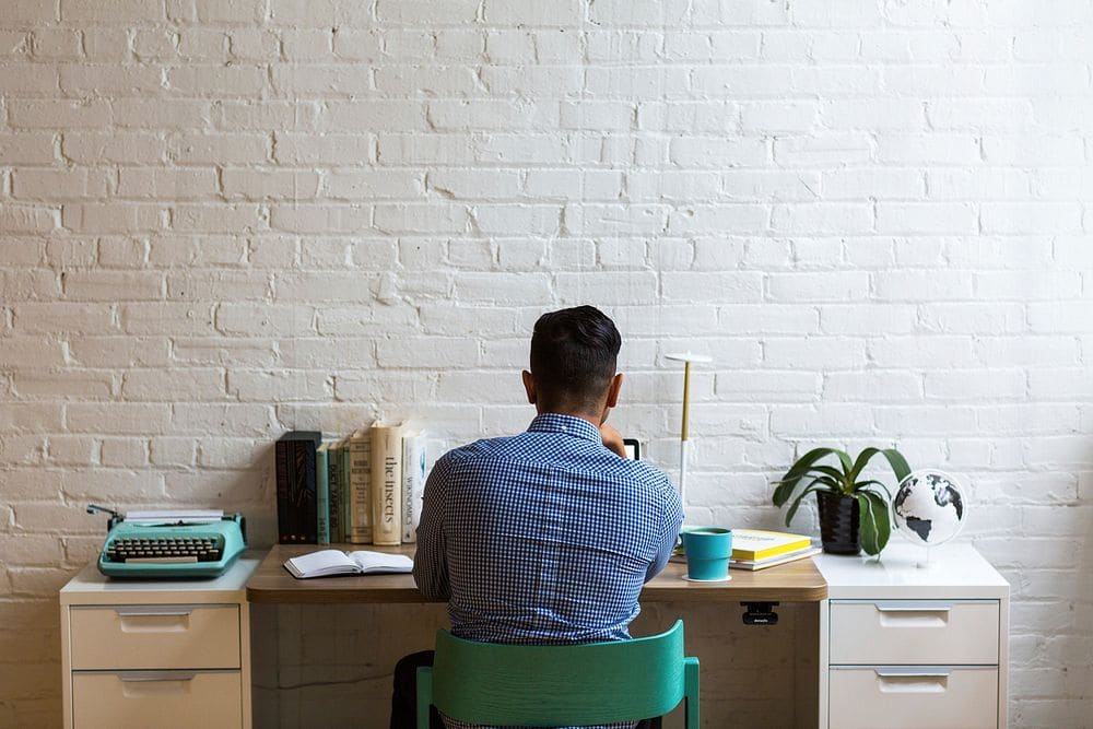 A man in a blue checked shirt working at a desk against a white brick wall, a small plant and books beside him.