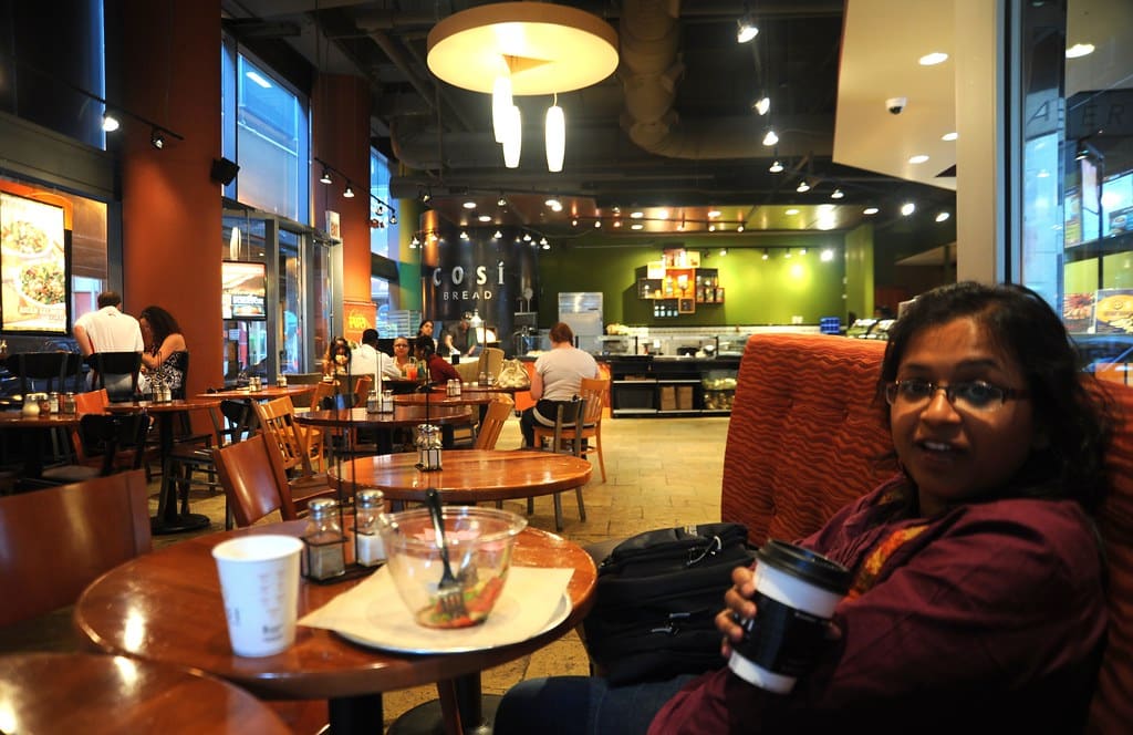 The warm interior of a busy café, with round tables, pendant lights and a takeaway cup in the foreground.