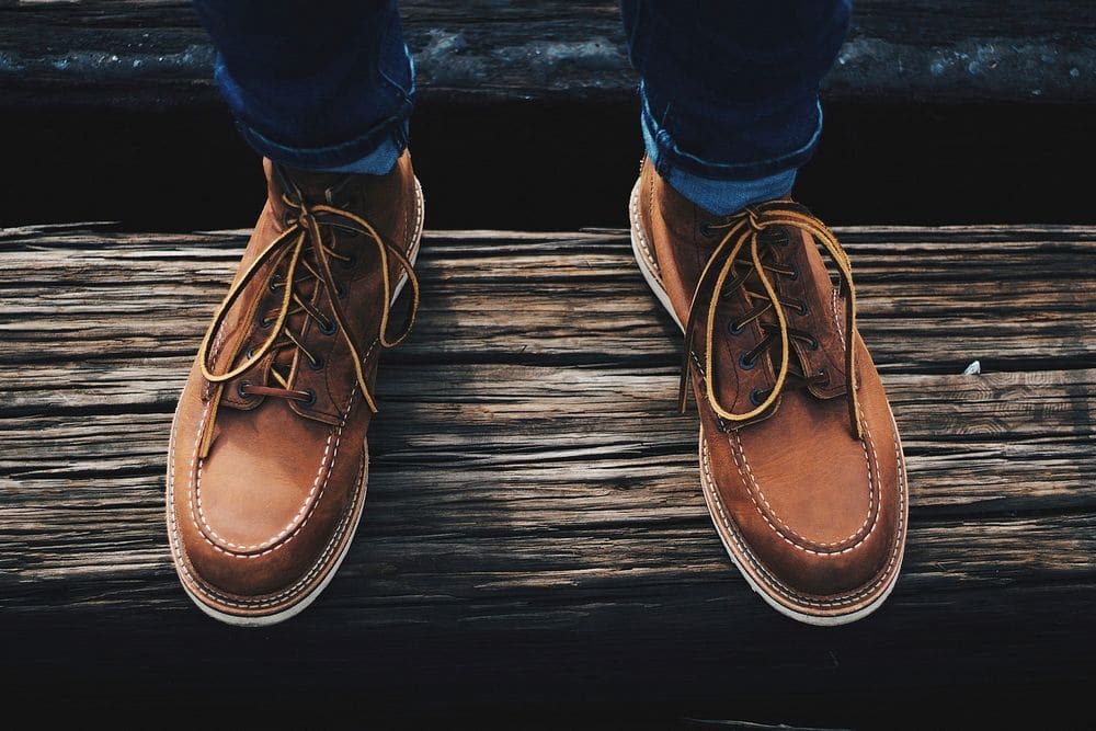 A pair of tan leather moc-toe work boots with yellow-flecked laces, worn with dark jeans and planted on a weathered wooden beam.