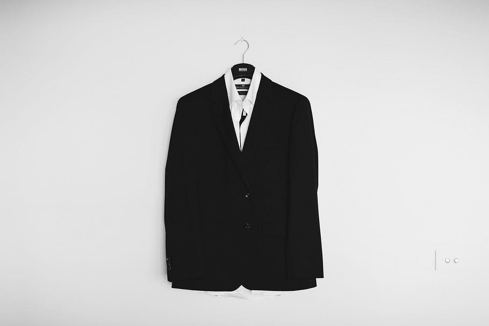 A black two-button suit jacket over a white shirt on a hanger against a plain white wall.