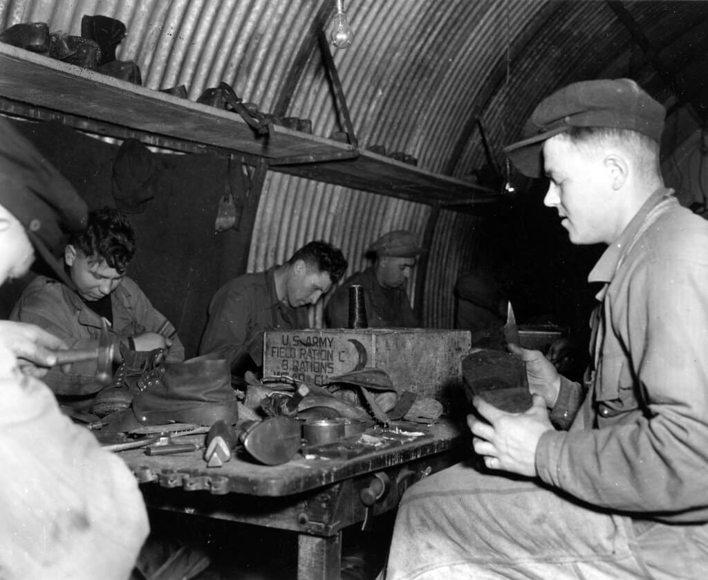 A black-and-white photograph of cobblers at a crowded workbench in a curved metal hut, one man holding a boot sole up to inspect it.