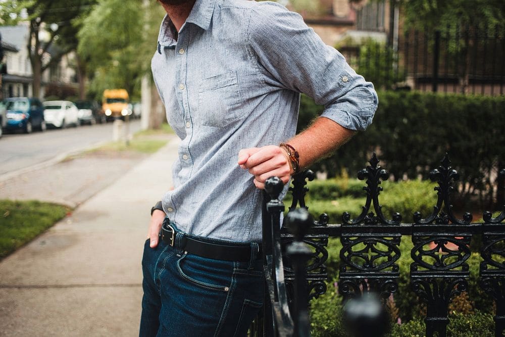 A man in a light blue button-down shirt with rolled sleeves and dark jeans, leaning on an iron fence on a leafy street.