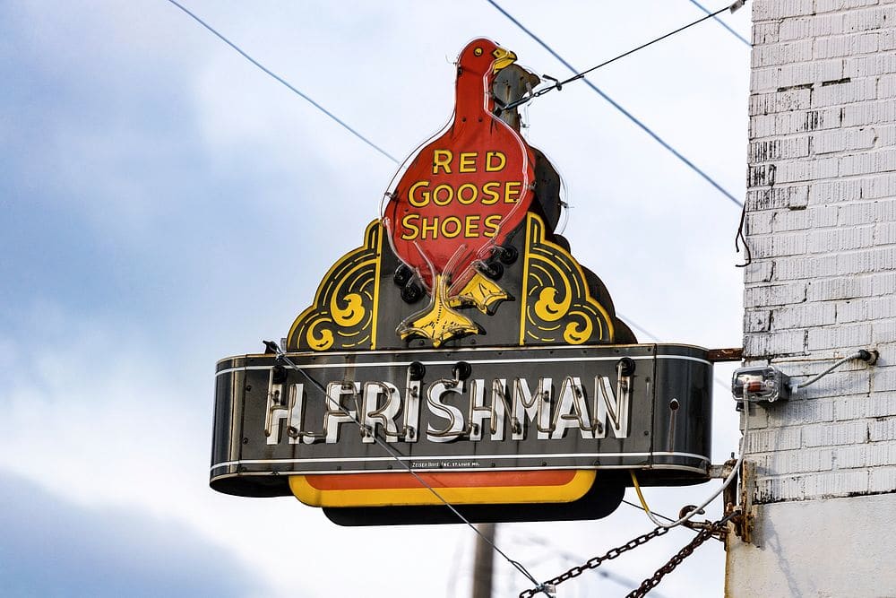 A vintage neon sign with a red goose and the words "Red Goose Shoes" above a shopkeeper's name, against a pale sky.