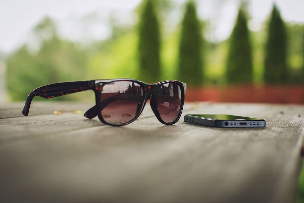 A pair of tortoiseshell sunglasses beside a smartphone on a wooden ledge, trees blurred behind.