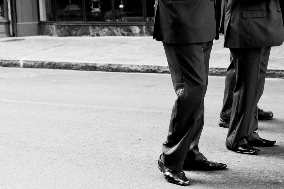 A black-and-white street shot of a man in dark suit trousers and polished black shoes mid-stride on a sidewalk.