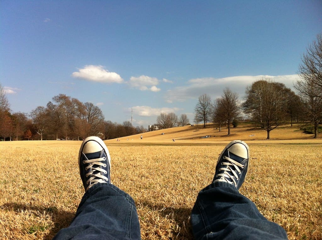 A first-person view of black canvas high-top sneakers and dark jeans stretched out on a dry grass field under a blue sky.