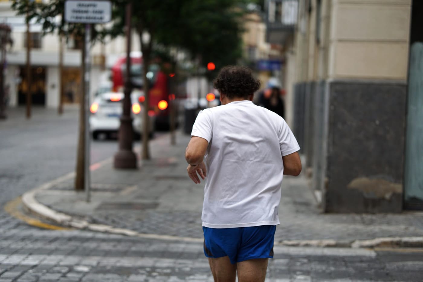A man in a plain white crewneck T-shirt and blue shorts jogging away down a city sidewalk.