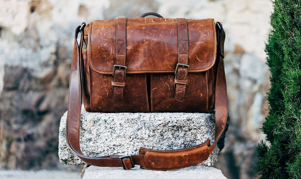 A brown leather messenger bag with two buckled straps and a shoulder strap, set on a granite block.