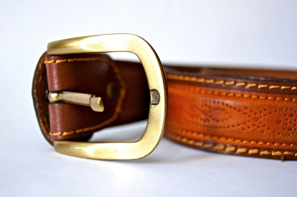 A tan leather belt with stitched edges and a solid brass buckle, shown close up.